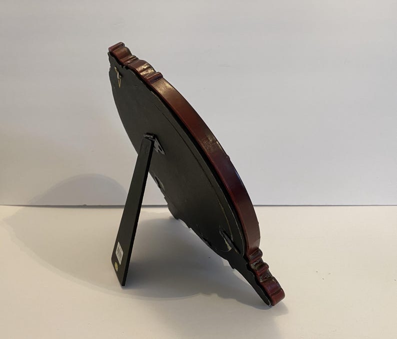 May include: A dark brown wood picture frame with a black backing and a decorative ornate design. The frame has a stand on the back to prop it up.