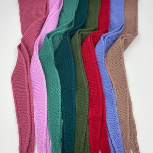 May include: A collection of eight long, narrow knitted scarves arranged side-by-side in a vertical orientation. The scarves feature a consistent garter stitch texture and are presented in a spectrum of colors including mauve, pink, teal, dark green, olive, red, periwinkle, and tan.