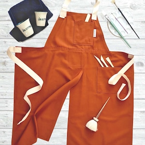 Może przedstawiać: Pomarańczowy fartuch z kremowymi paskami i kieszeniami, wraz z materiałami artystycznymi. Fartuch ma kieszeń z narzędziami i kieszeń na piersi. Obecne są również dwa małe ceramiczne kubki z napisem "deanna".