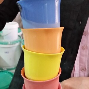 May include: A stack of five ceramic cups in different colors. The cups are stacked from largest to smallest, with the largest cup being light blue, followed by blue, orange, yellow, and pink.