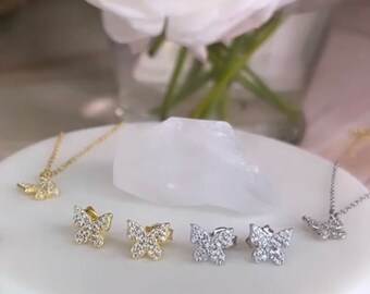 May include: A set of butterfly earrings and a butterfly necklace. The earrings are available in gold and silver, and the necklace is silver. All three pieces are adorned with sparkling clear stones.