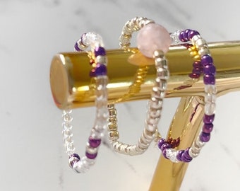May include: Three beaded rings on a gold stand. One ring is silver with white beads, one is silver with white and purple beads, and one is silver with pink beads.