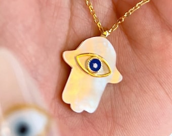 May include: A gold chain necklace with a white hamsa hand pendant featuring a blue evil eye charm. The pendant is made of mother of pearl.