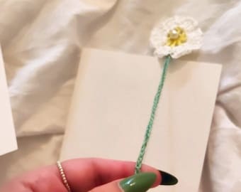 May include: A green and white crocheted flower bookmark with a yellow tassel. The bookmark is attached to a white card.