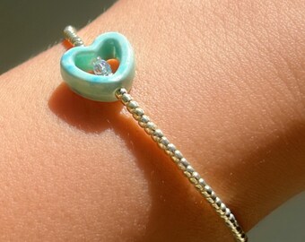 May include: A silver beaded bracelet with a light blue heart charm. The heart charm has a clear crystal in the centre.