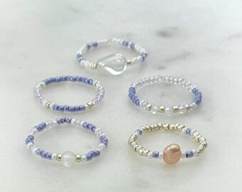 May include: A set of five beaded rings featuring small purple, white, and metallic silver seed beads. One ring includes a clear heart-shaped charm, another features a small white bead, and a third ring displays a single round peach-coloured bead.