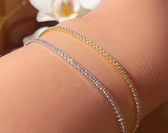 May include: Two delicate tennis bracelets, one in gold and one in silver, with a row of sparkling diamonds on each. The bracelets are worn on a person's wrist.