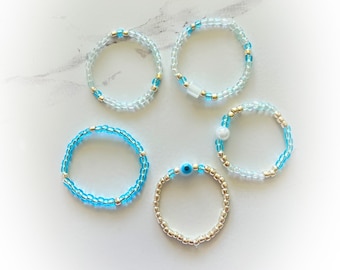May include: Five beaded bracelets with different charms. Two bracelets are light blue with gold beads, one has a white bead, one has a blue evil eye charm, and one has a light blue bead. The bracelets are on a white marble surface.