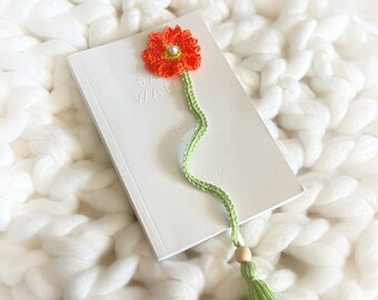 May include: A crocheted bookmark featuring a bright orange flower with a green tassel. The flower has a white pearl bead in the centre. The bookmark is on a white fluffy surface.