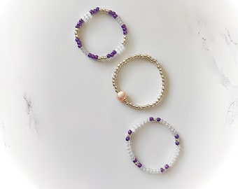May include: Three beaded bracelets with gold accents. One bracelet has a single pink pearl bead. Two bracelets have alternating white and purple beads.