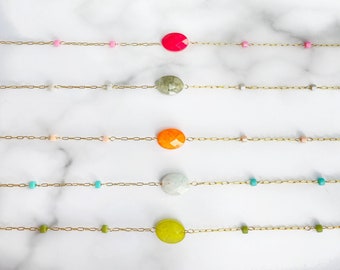 May include: Five gold chains with different coloured gemstone beads. Each chain has a different coloured gemstone bead, including pink, green, orange, turquoise, and yellow. The chains are laid out on a white marble surface. A green and white crescent moon shaped stone is in the top centre of the image.