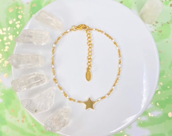 Puede incluir: Un brazalete de cadena dorada con un pequeño dije de estrella dorada. El brazalete está en un plato blanco con cristales de cuarzo transparentes.