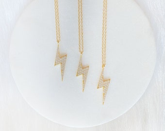 May include: Three gold necklaces with a lightning bolt pendant encrusted with small white stones. The necklaces are displayed on a white circular surface.