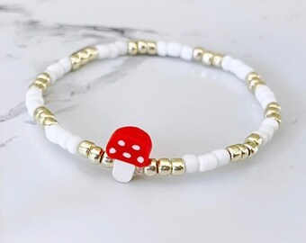 May include: A beaded bracelet with a red mushroom charm with white spots. The bracelet is made with white and gold-coloured beads, creating a whimsical accessory. The bracelet is a stretch style, suitable for various wrist sizes.