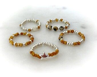 May include: Five beaded rings arranged in a circle. The rings feature a combination of metallic silver beads and tan-coloured beads. One ring includes a silver star charm, another features three spotted dalmatian jasper beads, and one has a white bead.