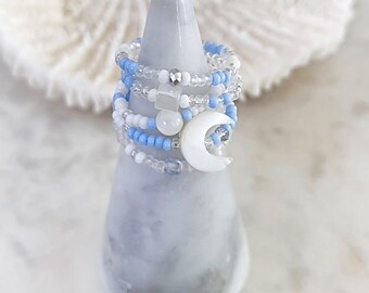 May include: A set of five beaded rings with a crescent moon charm. The rings are made with white and light blue beads and are stacked on a grey marble ring holder.