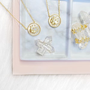 Puede incluir: Dos collares de oro con colgantes de luna y estrellas en una bandeja de m&aacute;rmol blanco con un cristal transparente. La bandeja est&aacute; sobre una superficie rosa.