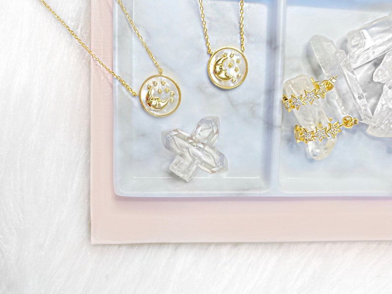 Puede incluir: Dos collares de oro con colgantes de luna y estrellas en una bandeja de m&aacute;rmol blanco con un cristal transparente. La bandeja est&aacute; sobre una superficie rosa.