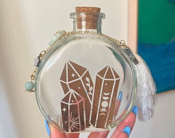 May include: A clear glass bottle with a cork stopper and a gold chain. The bottle features a gold design of crystals and celestial symbols. The bottle is decorated with small charms.