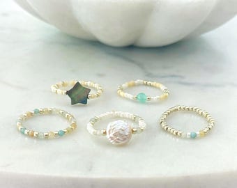 May include: A set of five beaded rings featuring small gold, white, and light blue glass beads. One ring displays a dark star-shaped charm, another features a central white textured circular bead, and a third includes a small light blue bead.