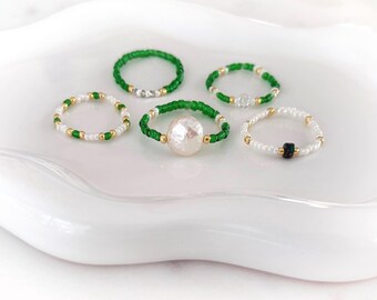 May include: A collection of five beaded rings featuring small green, white, and gold-toned beads. One ring is centred with a large, irregular white pearl-like bead, while another ring features a single small, dark green square-shaped bead.