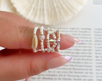 May include: A set of four beaded rings featuring white seed beads, a central tan oval bead, a clear round bead, and a central iridescent geometric crystal bead. The rings are arranged in a row, showcasing a variety of textures and bead shapes.