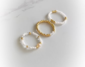May include: Set of four beaded rings with white and gold beads. The rings are stacked on top of each other.