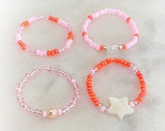 May include: Four beaded rings with different charms. The rings are made with pink and orange beads. One ring has a clear crystal charm, one has a pearl charm, and one has a white star charm.