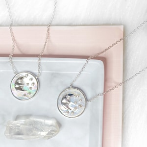 Puede incluir: Dos collares de plata con un colgante de luna creciente y estrella sobre un fondo blanco y rosa. El colgante est&aacute; hecho de n&aacute;car iridiscente con una luna creciente plateada y peque&ntilde;as estrellas plateadas.