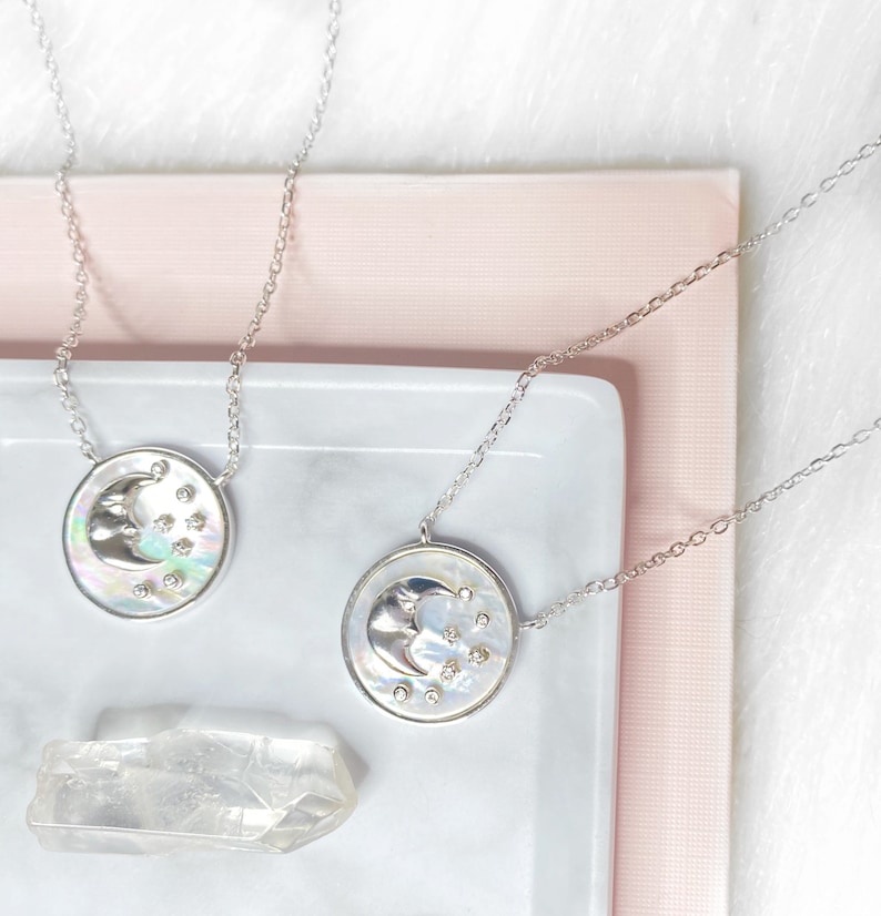 Puede incluir: Dos collares de plata con un colgante de luna creciente y estrella sobre un fondo blanco y rosa. El colgante est&aacute; hecho de n&aacute;car iridiscente con una luna creciente plateada y peque&ntilde;as estrellas plateadas.