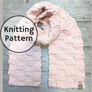 May include: A pink knitted scarf with a textured stitch pattern. The scarf is folded in half and laid flat on a wooden surface. The text "Knitting Pattern" is visible in a circle on the image.