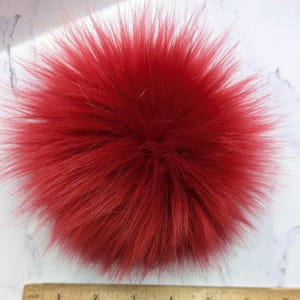 May include: A vibrant red faux fur pom-pom. The round pom-pom has long, fluffy strands radiating from the center. A wooden ruler with inch markings is at the bottom of the image.