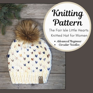 May include: A white knitted hat with a fair isle pattern of small hearts in blue, pink, and brown. The hat has a brown faux fur pom pom on top and a brown leather tag that says "Naptime Kst".