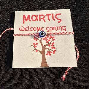 May include: A white card with the text "MARTIS" and "Welcome Spring" in red lettering. A red and white string with a blue evil eye charm is tied around the card. A brown tree with red leaves is printed on the card.