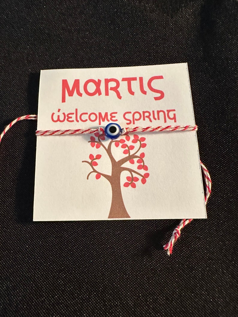 May include: A white card with the text "MARTIS" and "Welcome Spring" in red lettering. A red and white string with a blue evil eye charm is tied around the card. A brown tree with red leaves is printed on the card.