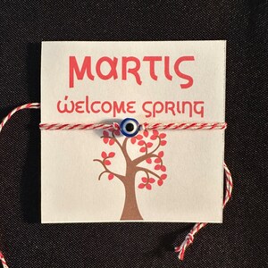 May include: A white card with red text that says "MARTIS" and "welcome spring". A red and white string is tied around the card with a blue evil eye charm in the centre. A brown tree with red leaves is printed on the card.
