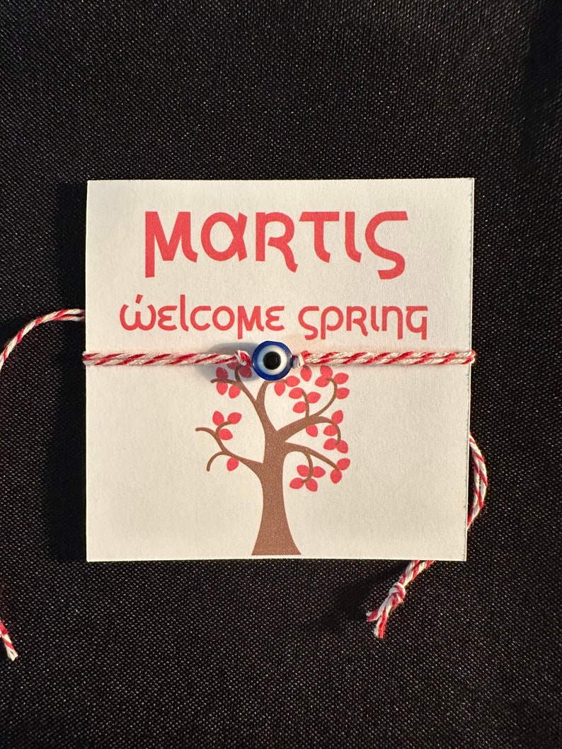 May include: A white card with red text that says "MARTIS" and "welcome spring". A red and white string is tied around the card with a blue evil eye charm in the centre. A brown tree with red leaves is printed on the card.