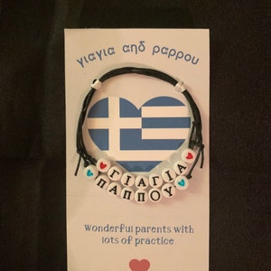 Peut inclure: Une carte blanche avec le texte "γιαγιά από παππού" et un bracelet en cordon noir avec des perles blanches qui épellent "γιαγιά παππού" et un cœur bleu et blanc avec le motif du drapeau grec. La carte dit également "Wonderful parents with lots of practice".