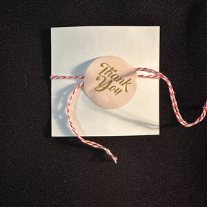 May include: A white card with a red and white string tied around it. A round sticker with the words "Thank You" in gold lettering is attached to the string.