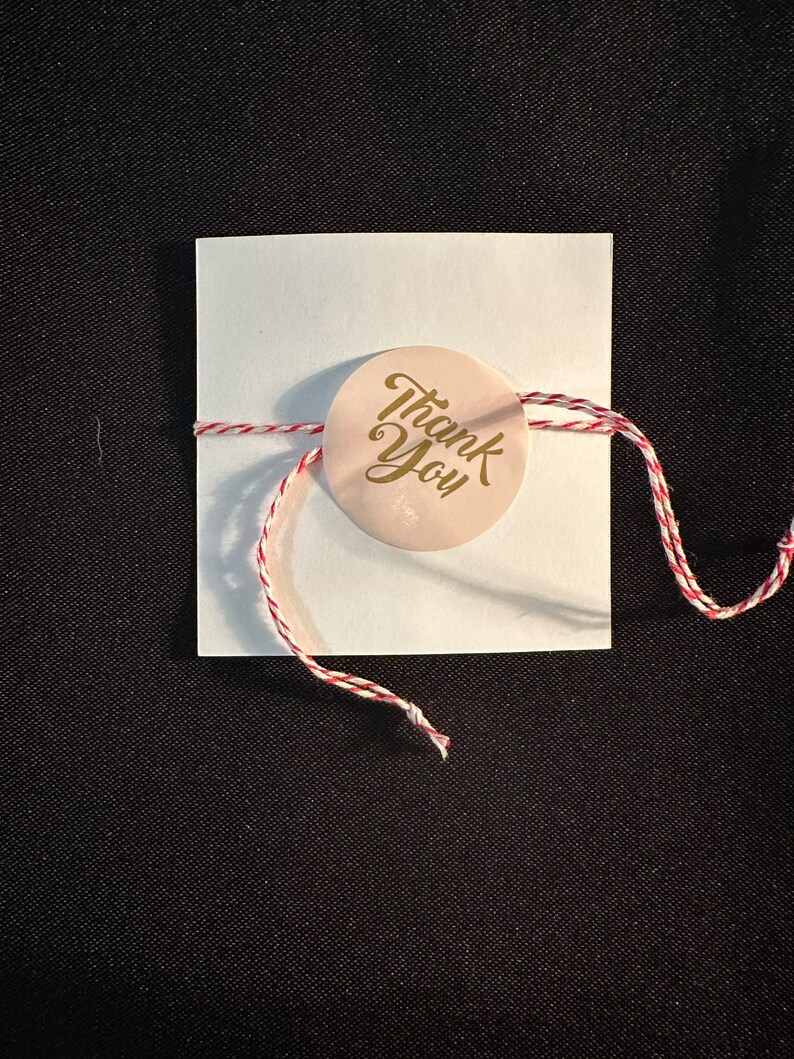 May include: A white card with a red and white string tied around it. A round sticker with the words "Thank You" in gold lettering is attached to the string.
