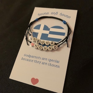 May include: A white card with blue and white Greek flag design and the text "nouna and nouno" in Greek. The card also has the text "Godparents are special because they are chosen" and a pink heart. A black string bracelet with silver beads spelling "NOUNA NOUNO" is on the card.