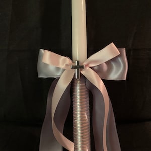 May include: A white candle with a silver cross, adorned with a pink and gray ribbon bow and streamers. The candle has a textured, silver-colored base. The background is black.