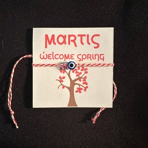 May include: A white card with the text "MARTIS" and "welcome spring" in red lettering. A red and white string with a blue evil eye charm is tied around the card. A brown tree with pink leaves is printed on the card.
