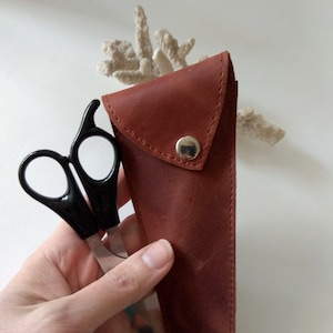 May include: A brown leather case with a snap closure, holding a pair of black handled scissors with silver blades.