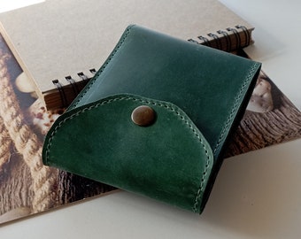 May include: A dark green leather wallet featuring a curved flap closure secured with a single metallic snap button. The wallet is constructed with visible stitching along the edges and has a smooth, matte finish on the exterior surface.