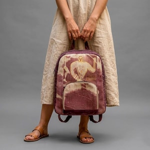 May include: A burgundy backpack with a front pocket and a zipper closure. The backpack features a beige and burgundy graphic print of a figure. The bag is held by a person wearing a beige dress and brown sandals.