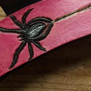 May include: Close-up of a pink leather item with a black spider design. The spider is intricately detailed, with a textured body and legs. A silver line runs through the center of the item, adding a contrasting element. The item appears to be a decorative piece.