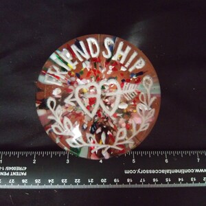 May include: A clear glass paperweight with a white design of two intertwined hearts with an arrow through them and the word "FRIENDSHIP" around the outside. The paperweight is filled with colorful confetti.