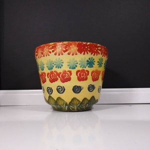 May include: A yellow ceramic planter with a colorful floral design. The planter has a wide rim and a rounded bottom. The design features red, blue, green, and yellow flowers.