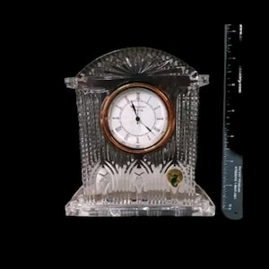 May include: A crystal clock with a gold-toned face and Roman numeral markings. The clock is in a clear crystal frame with a decorative design.